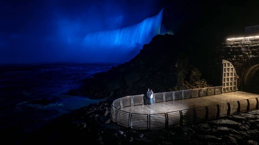 Couple standing on an observation deck at night, watching Niagara Falls illuminated in blue.